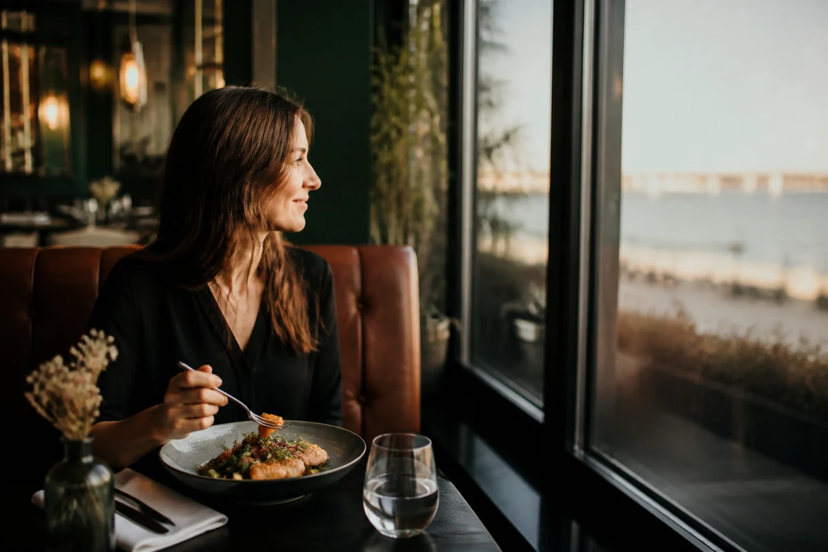 Femme attablée au restaurant devant une assiette de poisson et de légumes, regard vers la baie vitrée