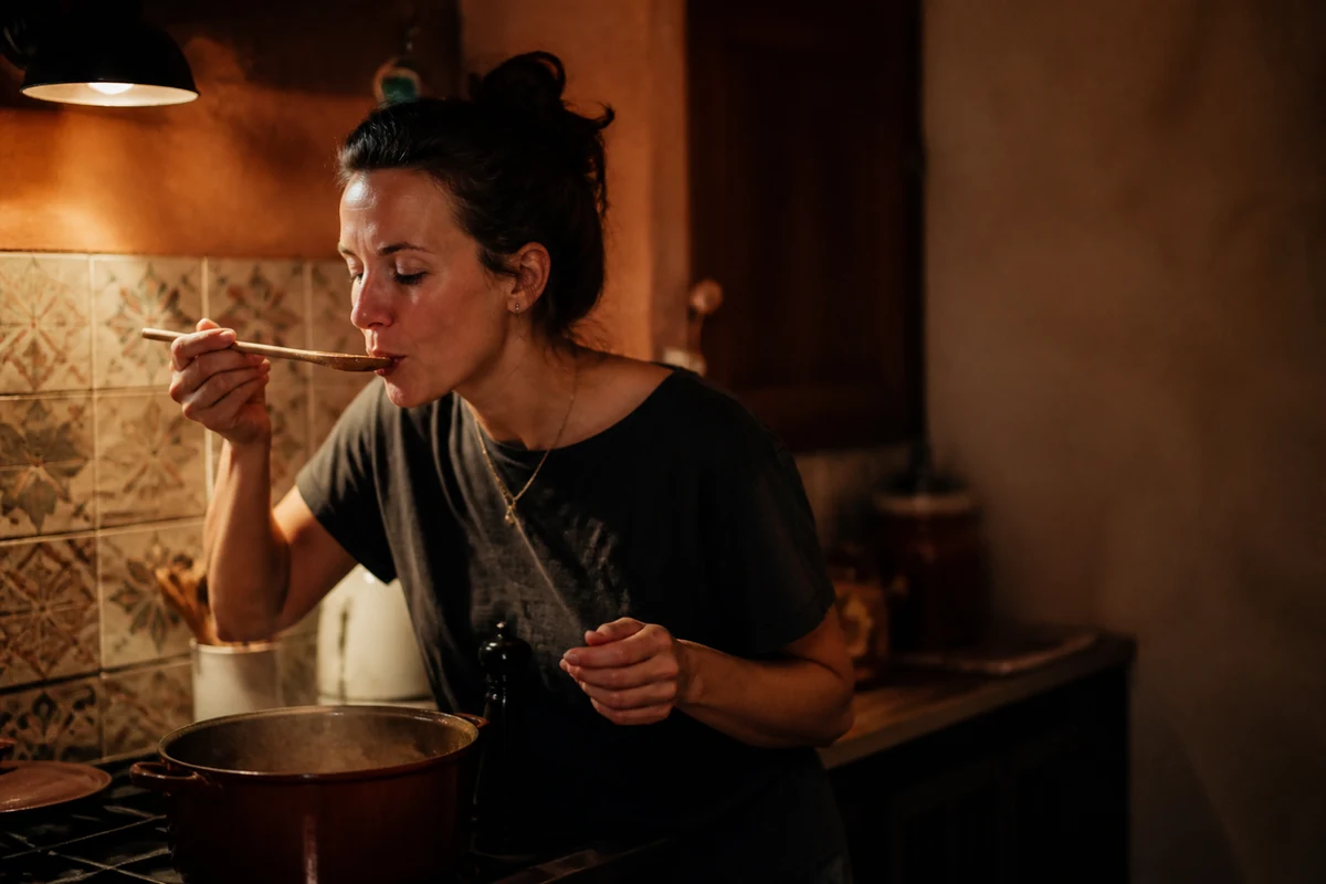 Femme goûtant une préparation à la cuillère en bois dans une cuisine chaleureuse en soirée